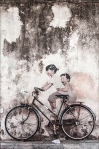 A realist street art wall mural of two life-size children positioned to appear as if they are riding an actual bicycle that is parked on a city sidewalk.